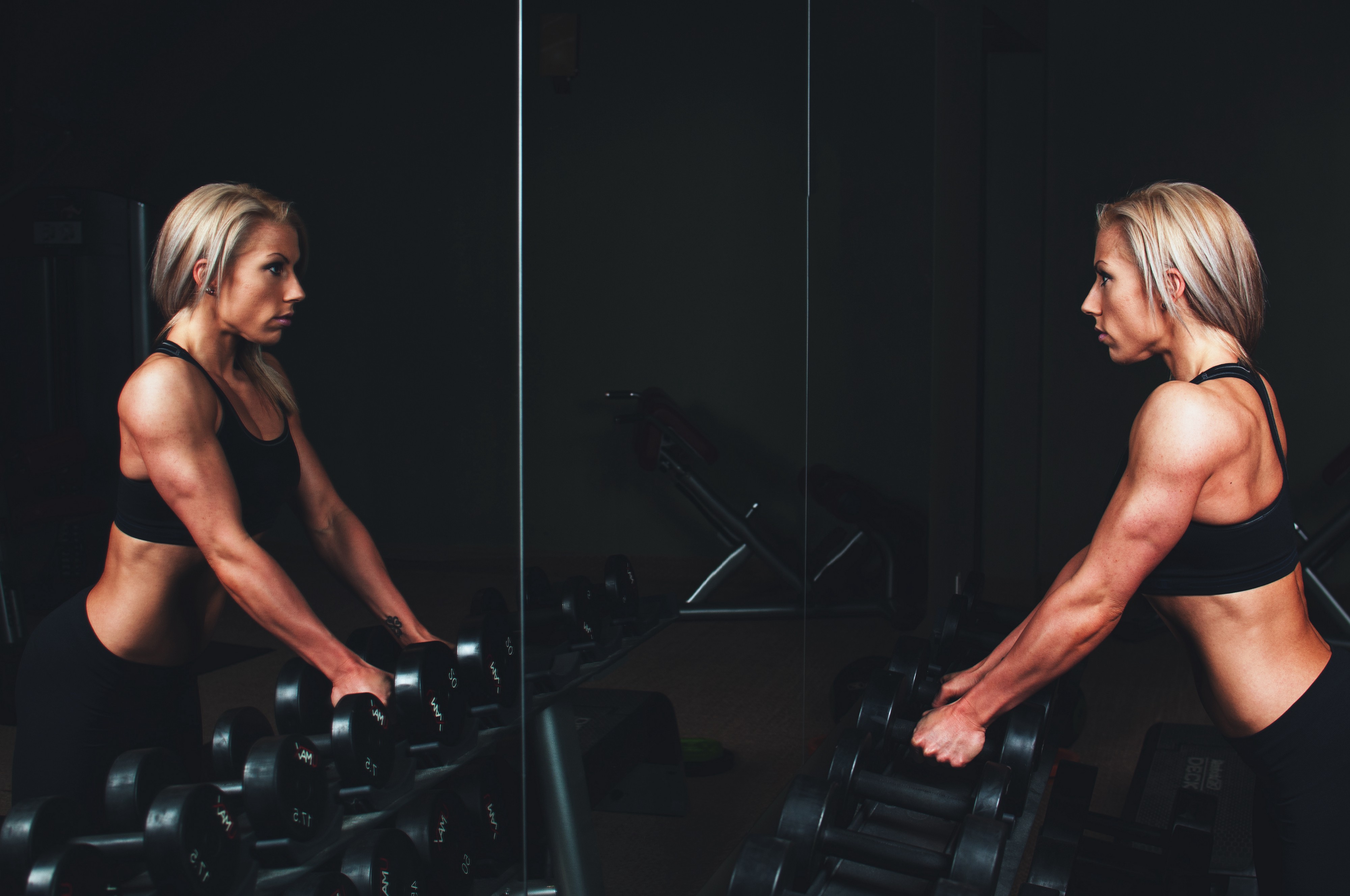 A very lean, fit woman leans against a dumbbell rack and stares at herself in a mirror.