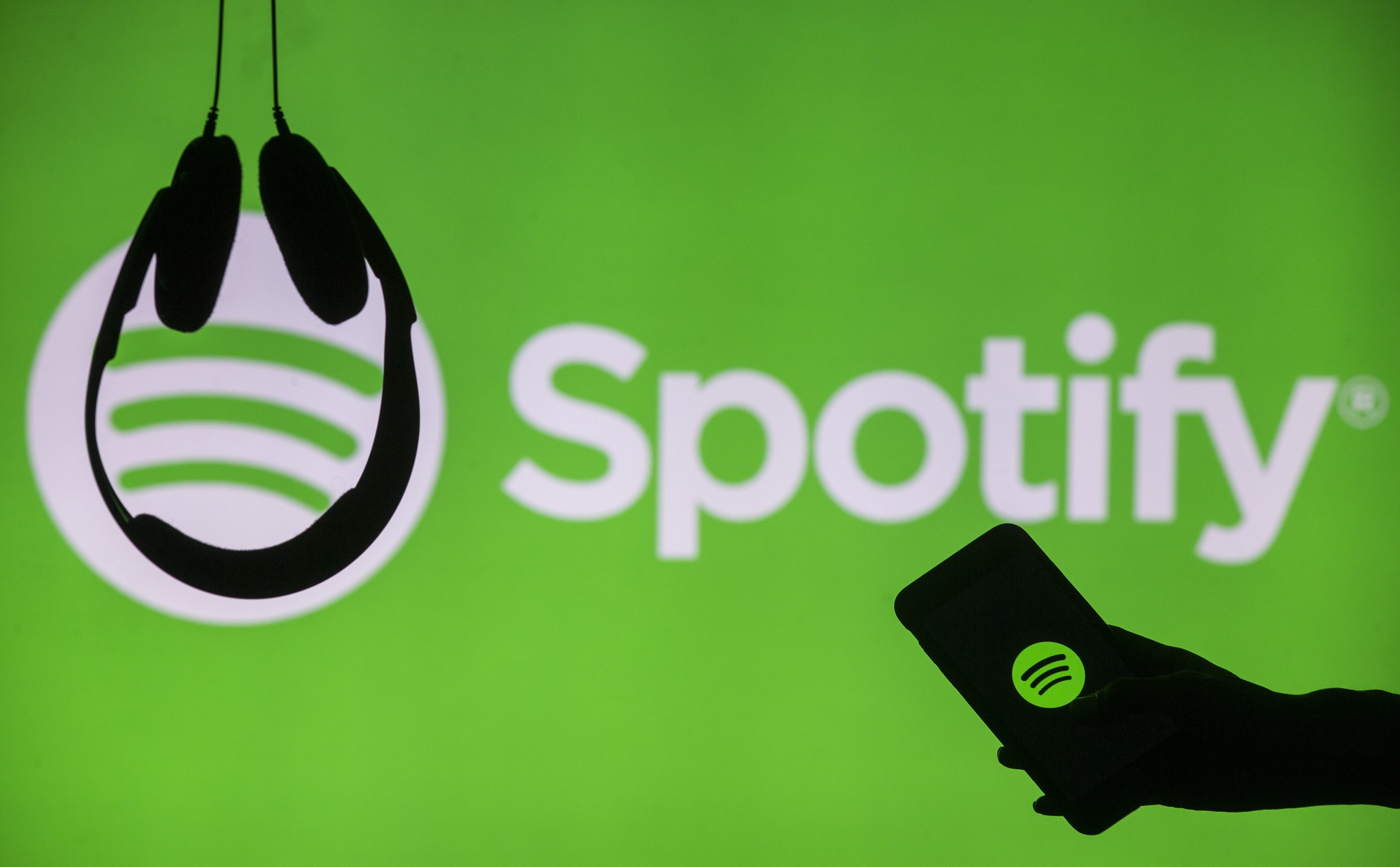 Headphones hang upside down as a hand holds a phone with the Spotify logo displayed in front of a screen showing Spotify