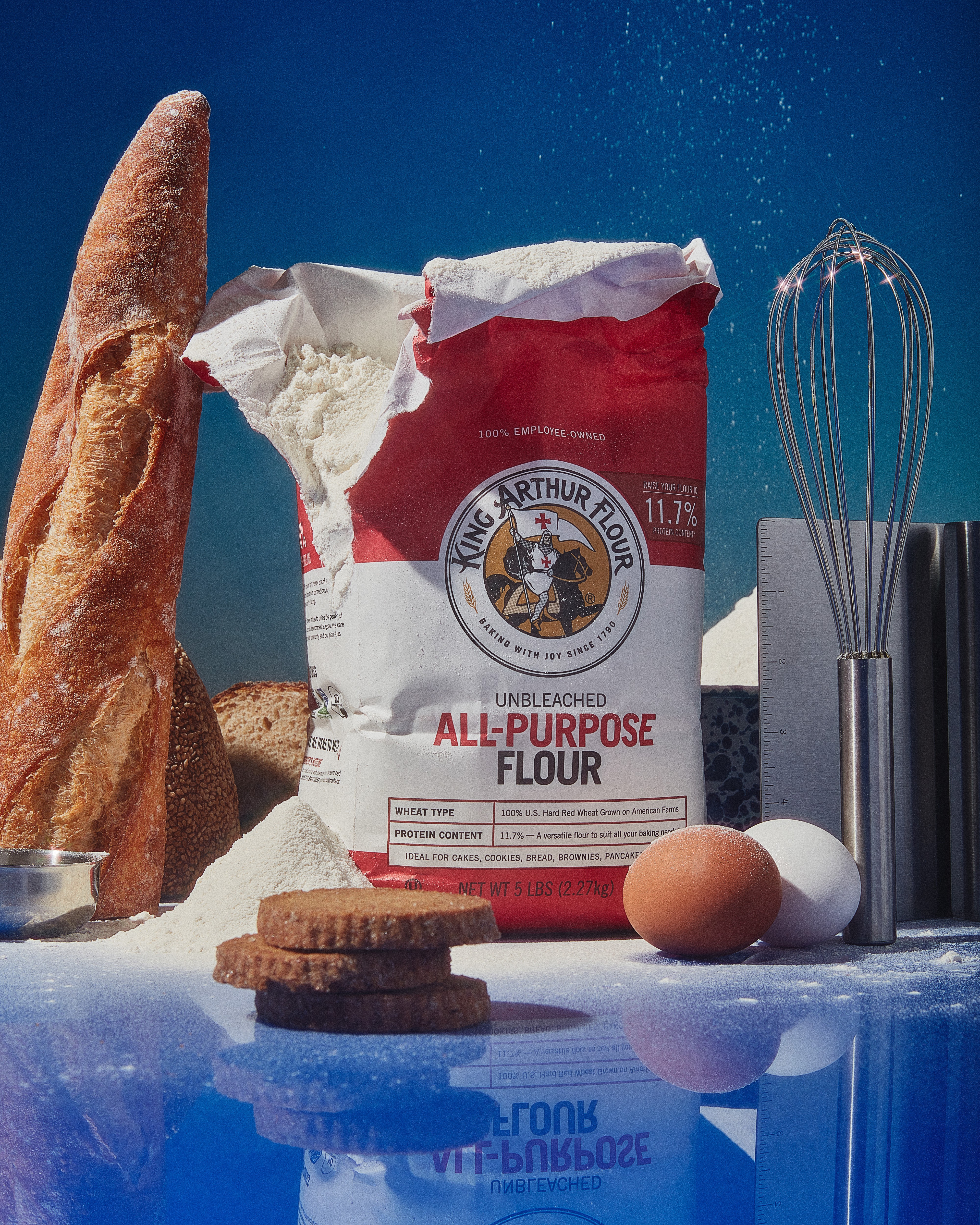 A bag of King Arthur Flour propped up next to a baguette, eggs, and a whisk.